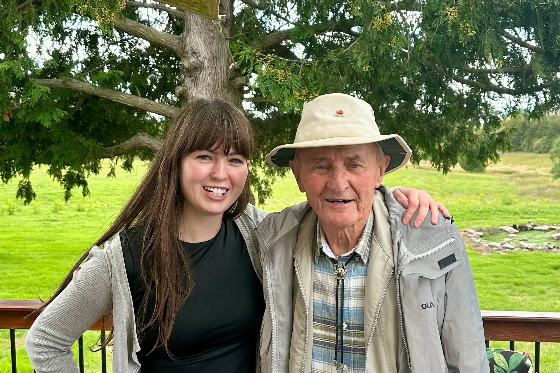 An elderly man and a young woman posing together on a wooden deck. The man is wearing a beige wide-brim hat, a light gray jacket over a blue and white plaid shirt, and khaki pants.