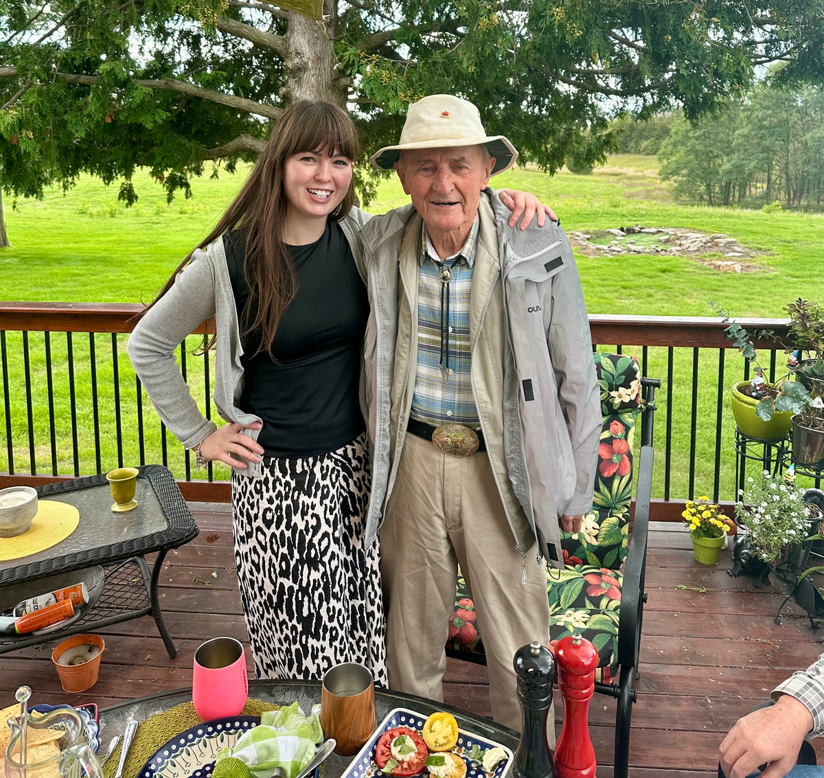 An elderly man and a young woman posing together on a wooden deck. The man is wearing a beige wide-brim hat, a light gray jacket over a blue and white plaid shirt, and khaki pants.