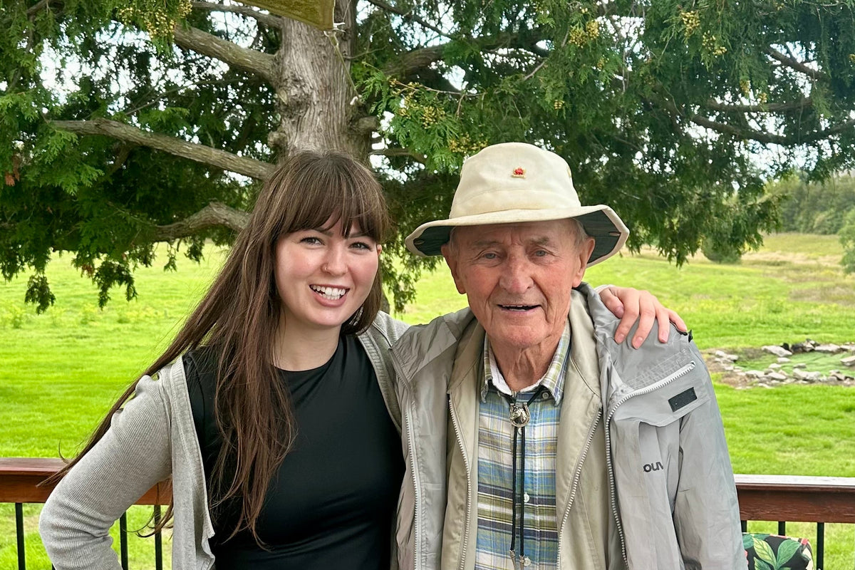 An elderly man and a young woman posing together on a wooden deck. The man is wearing a beige wide-brim hat, a light gray jacket over a blue and white plaid shirt, and khaki pants.