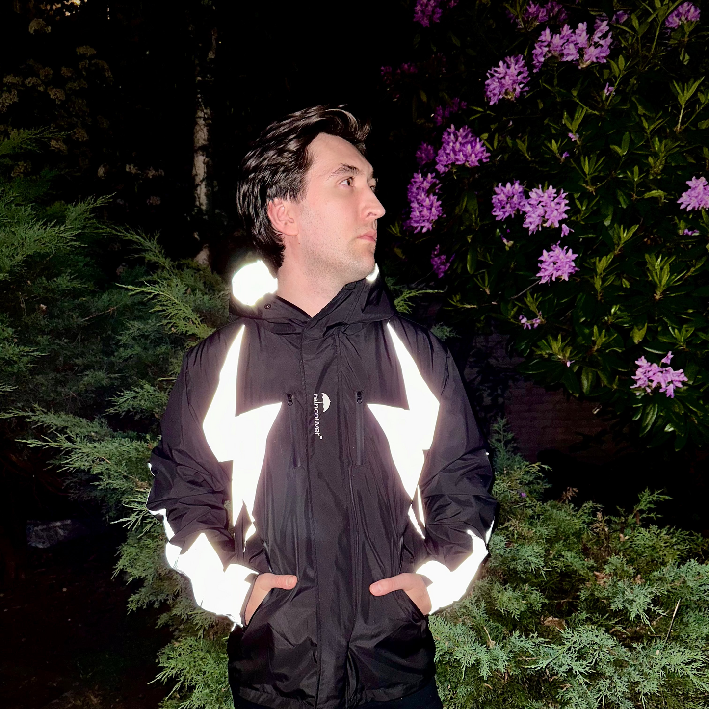 Person wearing a reflective black jacket standing outdoors with greenery and purple flowers in the background